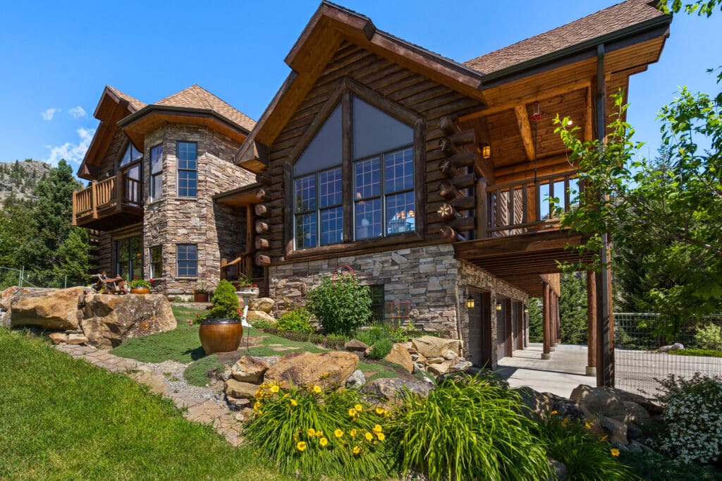 A large rustic log cabin with stone accents, large windows, and a sloped roof sits on a landscaped hill with yellow flowers—an ideal hunting property or cattle ranch under a clear blue sky.