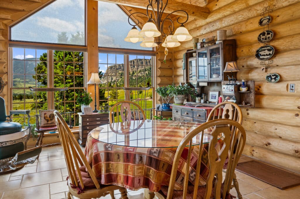 A cozy log cabin dining area with a round table and four chairs overlooks scenic mountains and greenery—ideal for those seeking recreational land or a hunting property; warm wood tones and rustic decor create an inviting atmosphere.