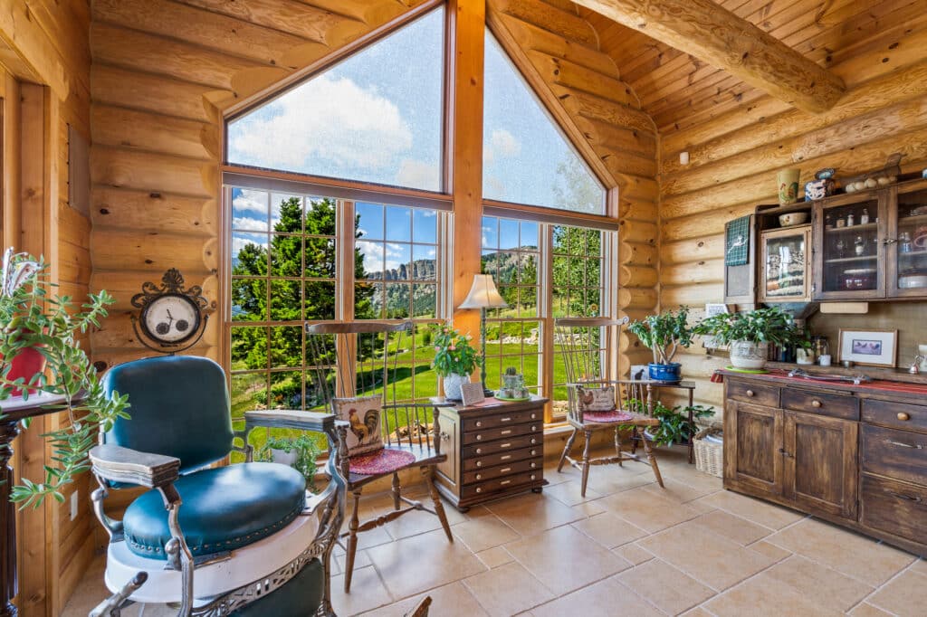 A cozy log cabin room with large windows overlooking recreational land, wooden walls and ceiling, potted plants, vintage chairs, a lamp, and rustic wooden furniture filled with decor.