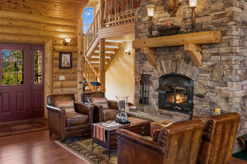 A cozy log cabin living room with leather chairs, a stone fireplace, wood walls and ceiling, a staircase, and sunlight streaming through large windows near a red front door—perfect for a hunting property or cattle ranch.