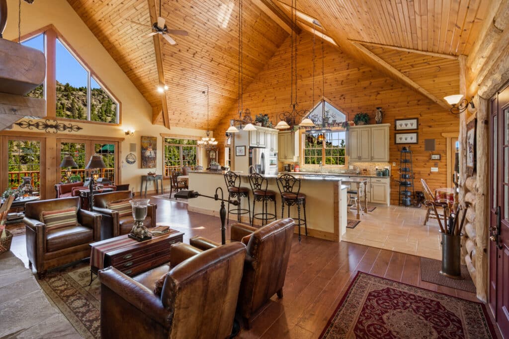 A spacious rustic living room and kitchen perfect for a hunting property, with high vaulted wooden ceilings, large windows, leather chairs, a marble island with barstools, and wooden floors that let in natural light and views of greenery outside.