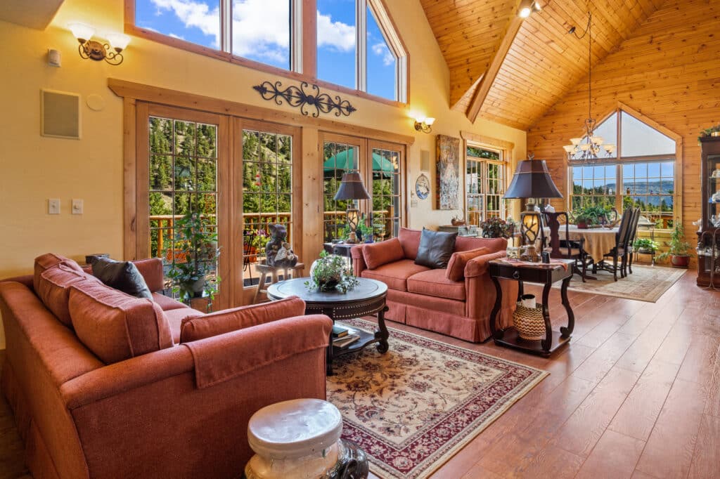 Spacious living and dining area with large windows, high wood-paneled ceiling, red sofas, decorative rug, indoor plants, and views of trees and blue sky—perfect for relaxing after exploring your ranch for sale or recreational land.