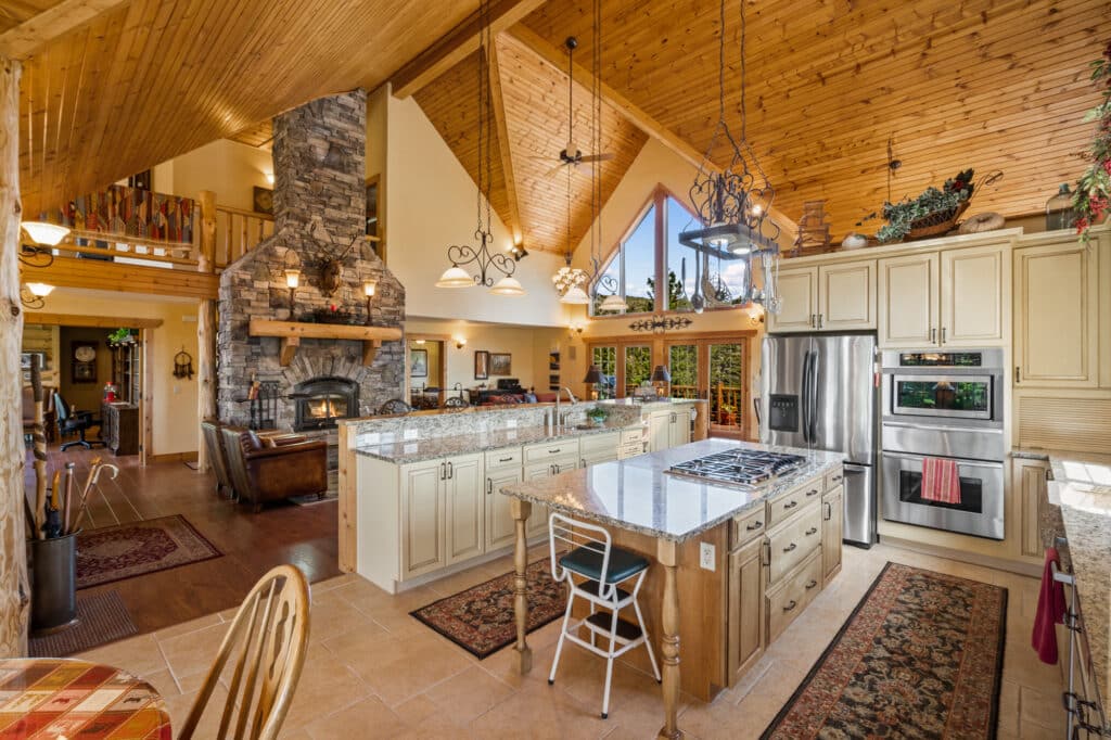 Spacious rustic kitchen with wood-paneled vaulted ceiling, stone fireplace, large island with cooktop, modern appliances, beige cabinets, granite counters—ideal for a cattle ranch or hunting property—with open views to a cozy living area and loft above.