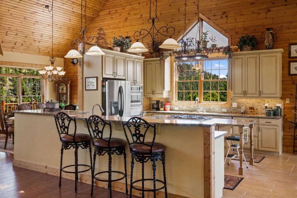 Spacious kitchen with cream cabinets, granite counters, a central island with ornate barstools, stainless steel appliances, hanging lights, and large windows overlooking green trees. Perfect for a cattle ranch or land for sale. Wood-paneled walls and ceiling add warmth.