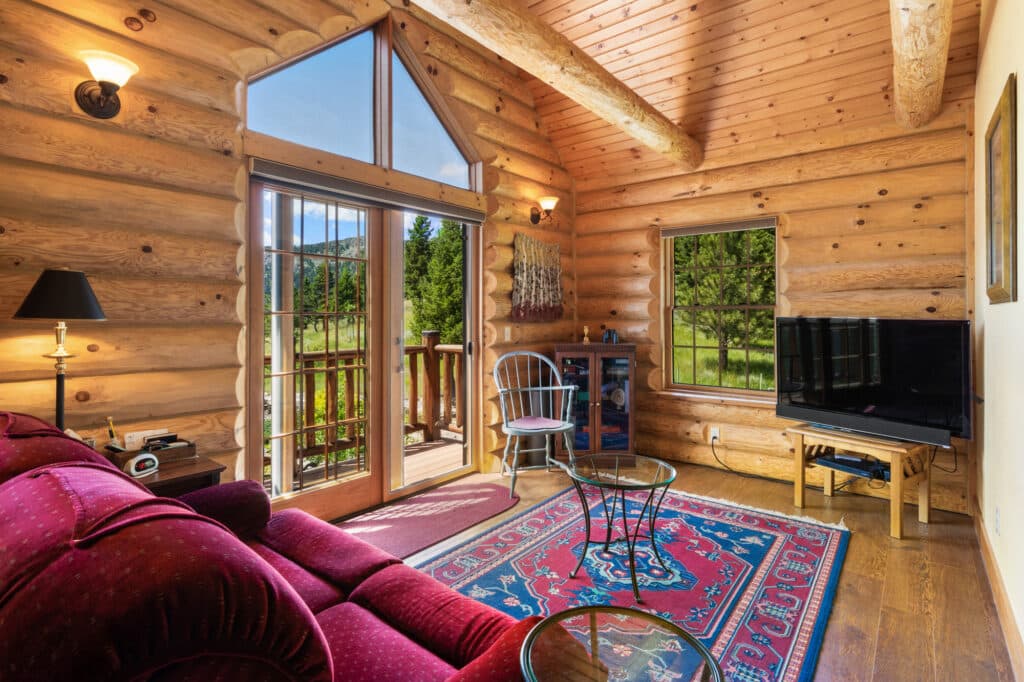Cozy log cabin living room with a red sofa, colorful rug, TV, and glass doors opening to a wooden porch. Sunlight streams in, highlighting the wood walls and peaceful forest view—ideal for those seeking ranch for sale or hunting property.