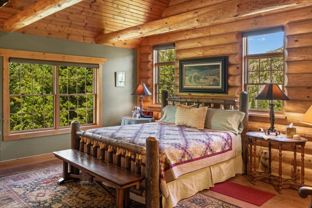 A cozy log cabin bedroom with a large bed, quilted bedspread, wooden furniture, two lamps on round side tables, a rug, and large windows overlooking green trees and recreational land. Warm, natural light fills the room.