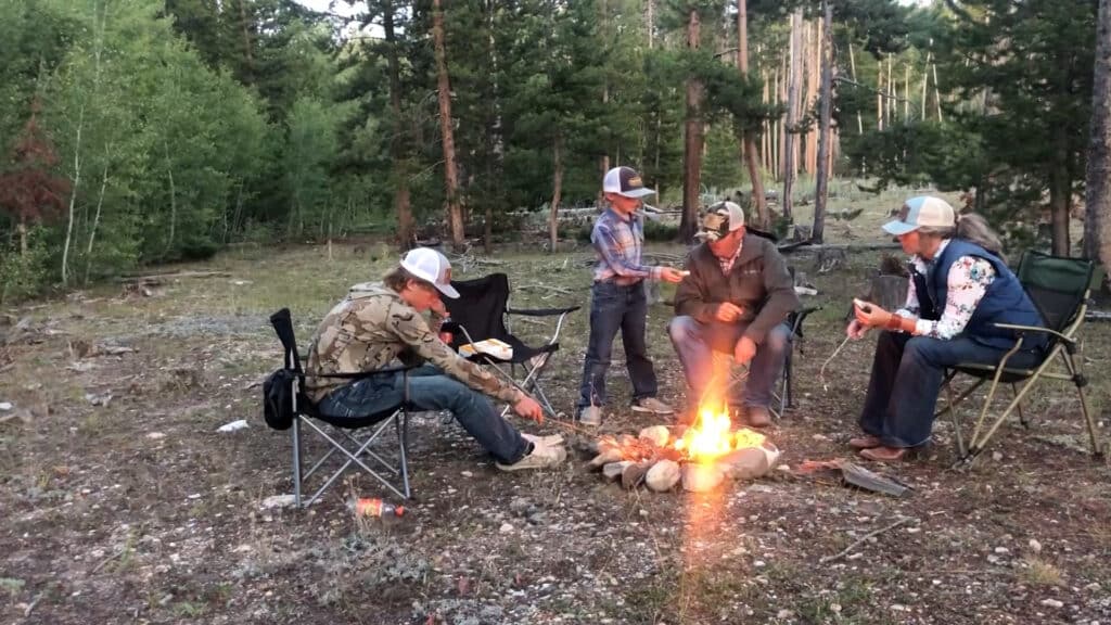Four people in outdoor clothing sit around a campfire in a forest clearing on recreational land, roasting marshmallows. Two adults and two children are present, with trees and greenery creating a peaceful backdrop.