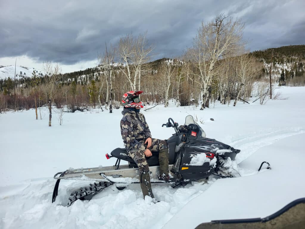 A person in camouflage winter gear and a helmet sits on a snowmobile in a snowy landscape with leafless trees and mountains, perfect for exploring recreational land or discovering your ideal ranch for sale.