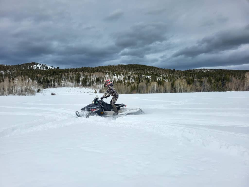 A person wearing a helmet rides a snowmobile across a wide snowy field on recreational land, with forested hills and cloudy skies in the background.