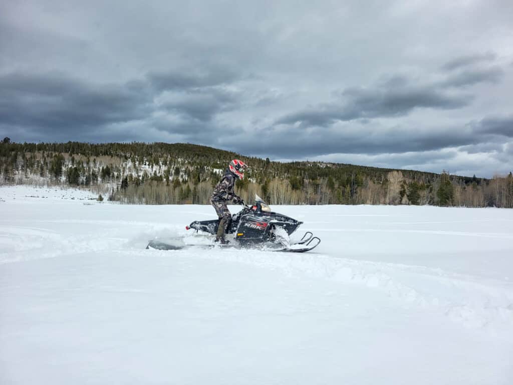 A person wearing a helmet and winter gear rides a snowmobile across a snowy field on recreational land, with a forest and cloudy sky in the background.