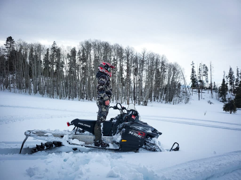 A person in camouflaged winter gear and helmet stands on a black snowmobile in a snowy landscape, with tracks in the snow trailing through a forest of leafless trees—a perfect scene for exploring recreational land or hunting property.