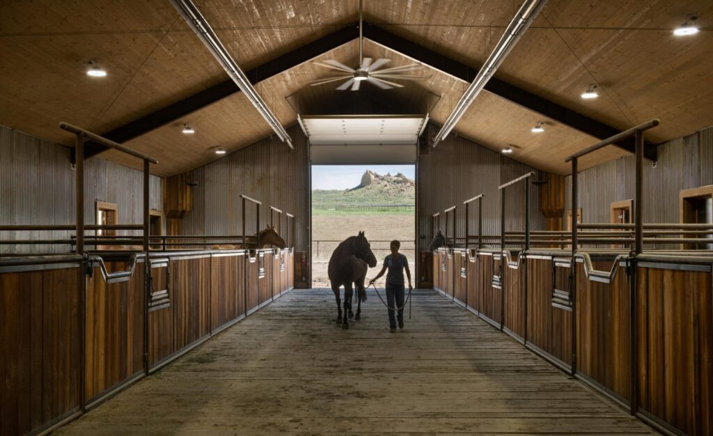 A person leads a horse down the center aisle of a wooden stable on a cattle ranch, with stalls on each side and daylight streaming in from an open door at the far end, revealing scenic ranch for sale views outside.