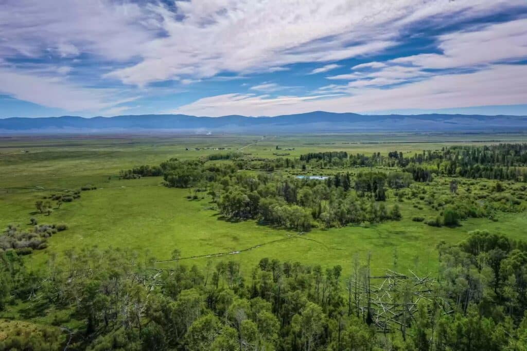 Aerial view of a vast green landscape with scattered trees, open fields, a small pond, and distant mountains under a partly cloudy sky—ideal as recreational land or hunting property.