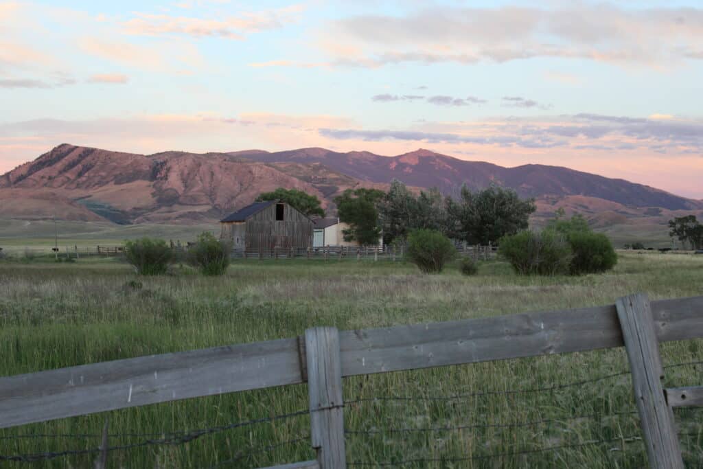 A rustic wooden barn sits in a grassy field, surrounded by trees and a wooden fence, with mountains in the background under a pink and blue sunset sky—ideal as a ranch for sale or hunting property.
