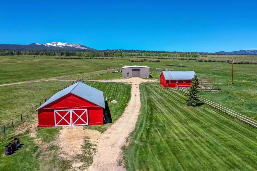 Aerial view of a rural ranch for sale with two red barns, a gray storage building, dirt driveway, green fields, and mountains with some snow in the background under a clear blue sky.