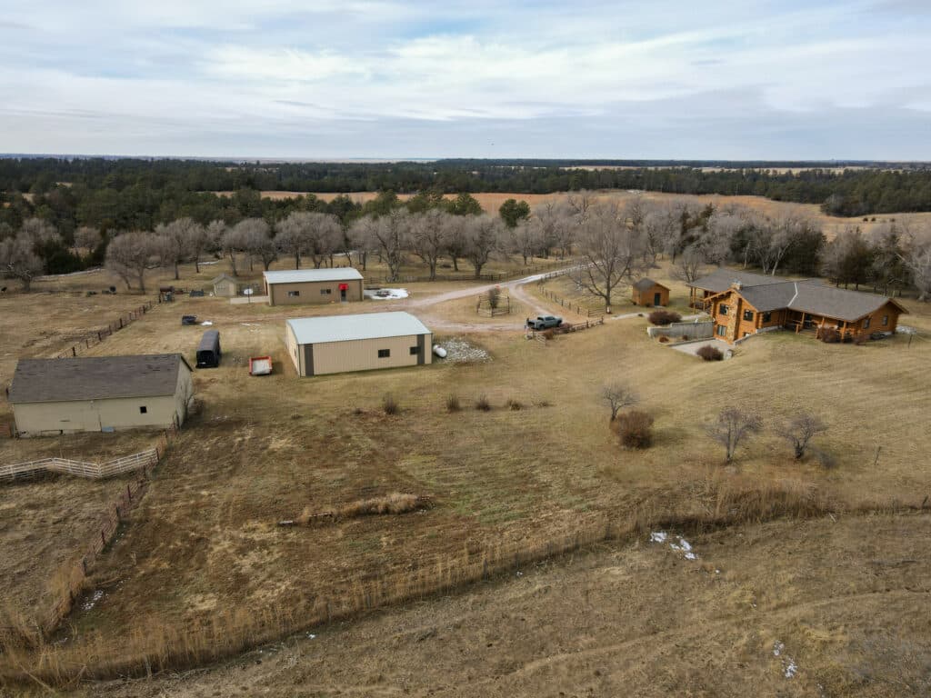 Aerial view of a rural property with several buildings, ideal as a hunting property or cattle ranch, surrounded by fields, trees, and a dirt driveway on a cloudy day.