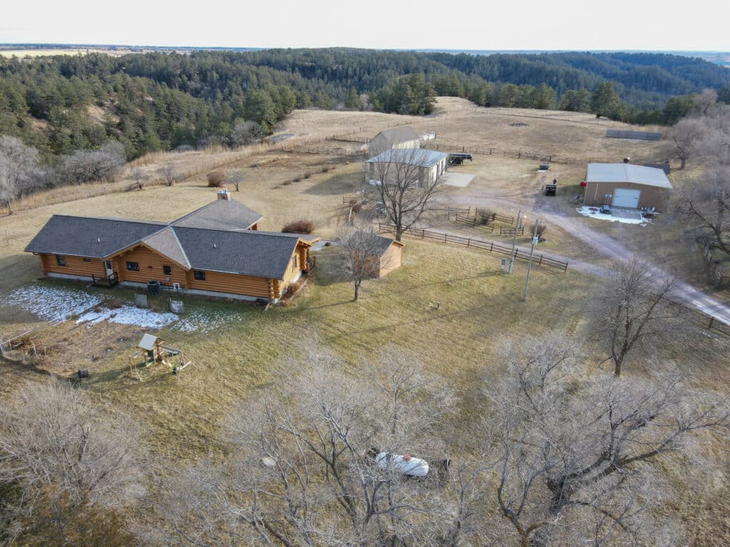 Aerial view of a rural hunting property with a wooden house, small barn, large shed, fenced areas, and surrounding grassland bordered by trees and hills. Some patches of snow are visible on this recreational land for sale.