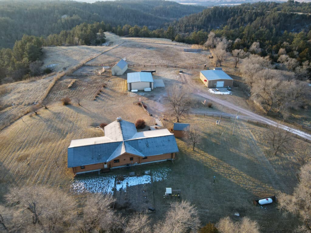 Aerial view of a rural hunting property with a large house, several outbuildings, patchy snow, open fields, trees, and a dirt driveway winding through the landscape. Ideal land for sale for those seeking privacy and outdoor adventure.