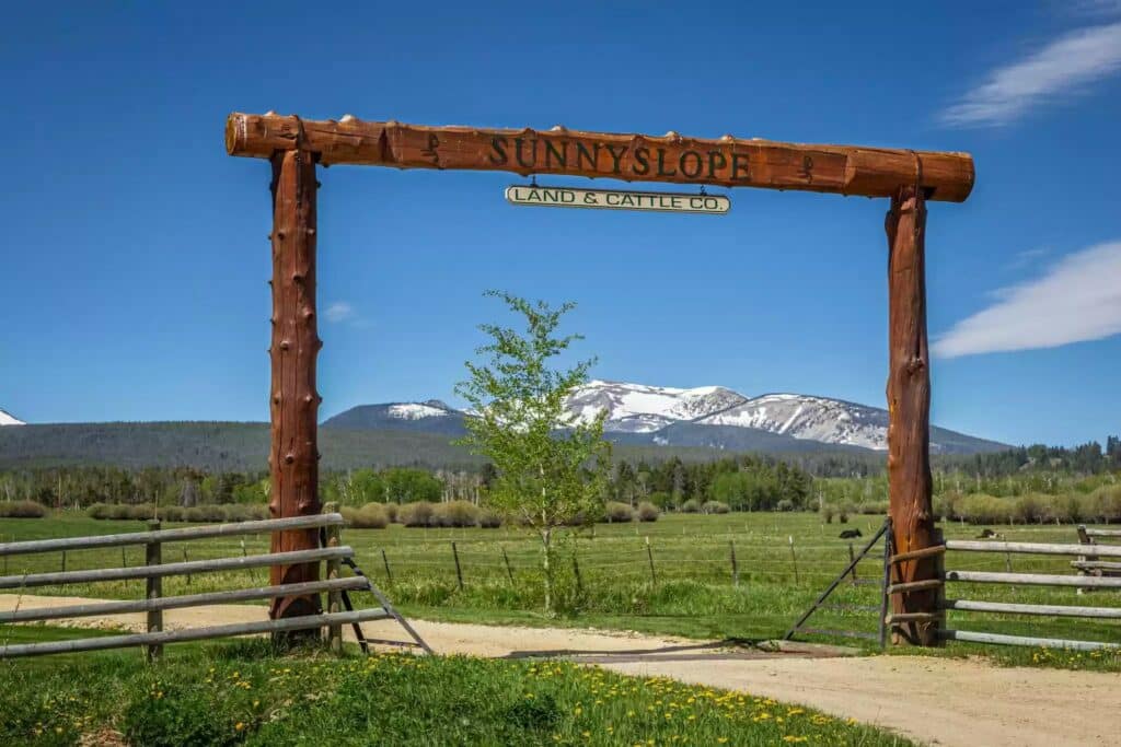 A wooden entrance sign reading “SUNNYSLOPE LAND & CATTLE CO.” stands over a dirt road, with green fields, a small tree, and snow-capped mountains beneath a clear blue sky—ideal land for sale or the perfect ranch for sale setting.