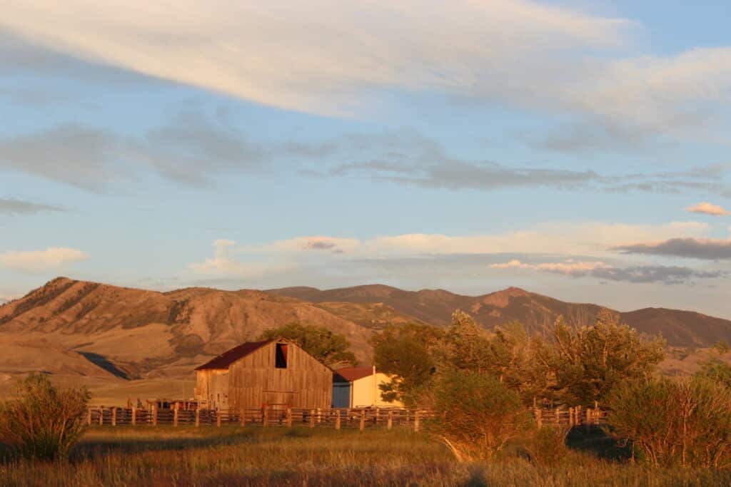 A wooden barn and small white building sit on recreational land in a grassy field, surrounded by trees and a wooden fence, with mountains and a partly cloudy sky in the background at sunset.