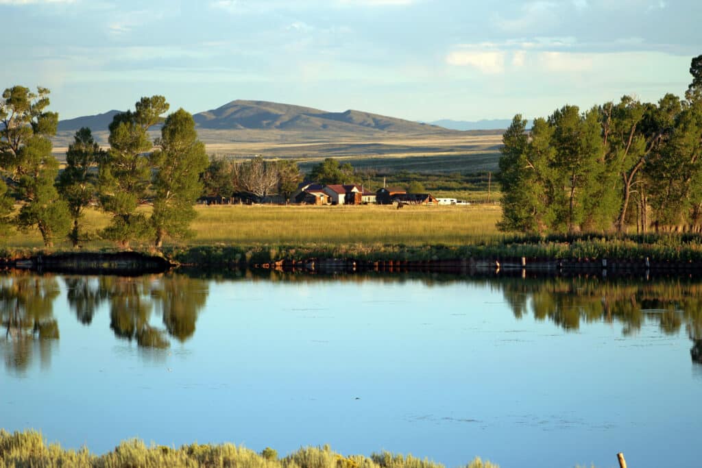 A calm lake reflects green trees and a distant farmhouse on recreational land, framed by mountains under a blue sky with scattered clouds.