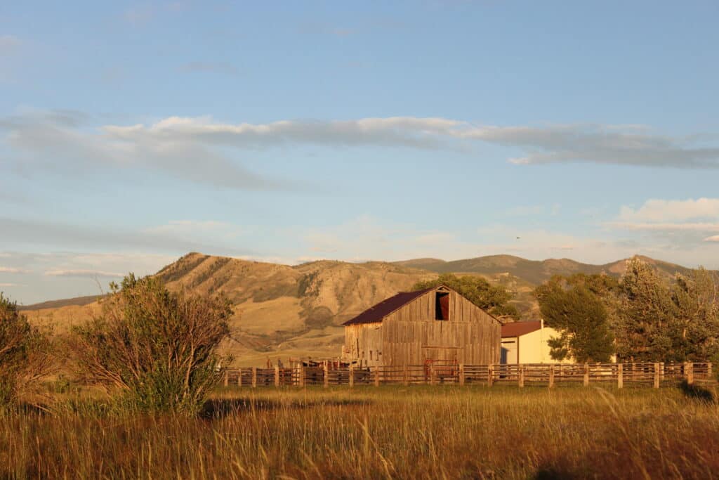 A rustic wooden barn and a small building sit in a grassy field on a cattle ranch, surrounded by trees and a wooden fence, with rolling hills and mountains in the background under a blue sky.