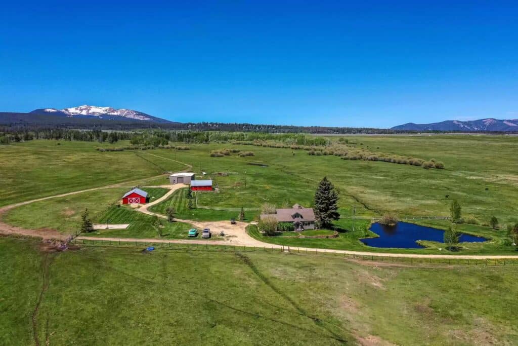 Aerial view of a rural cattle ranch with a house, pond, red barns, and several cars, set in a vast green field with mountains and a snow-capped peak in the background under a clear blue sky—prime land for sale.