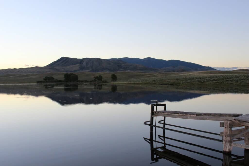 A wooden dock extends over a calm lake on recreational land, reflecting nearby hills and distant mountains under a clear sky at dusk.