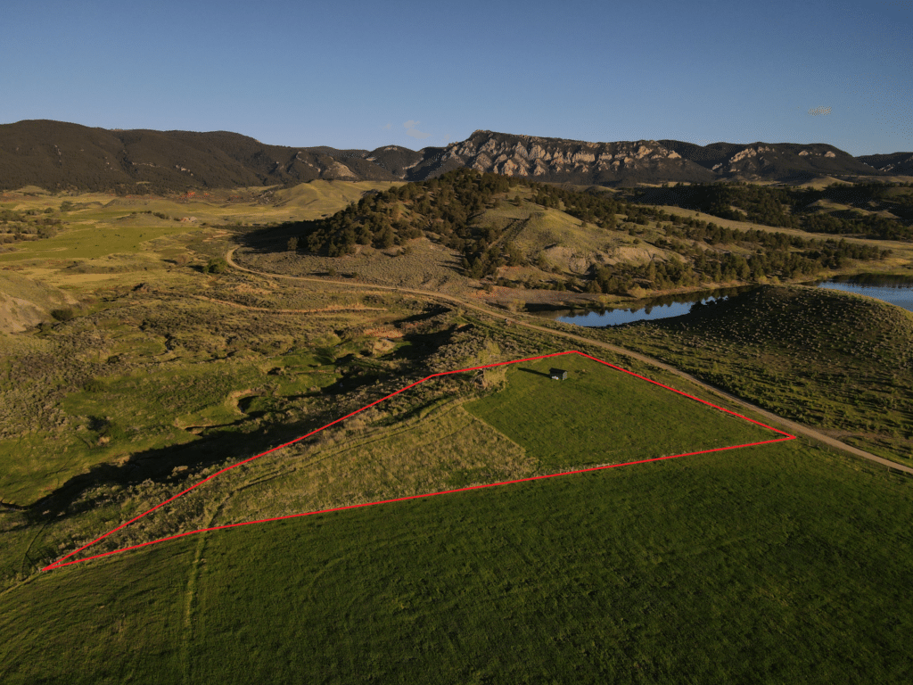 Aerial view of recreational land with a fenced area outlined in red, located near a body of water and rolling hills with rocky outcrops under a clear blue sky—an ideal ranch for sale.