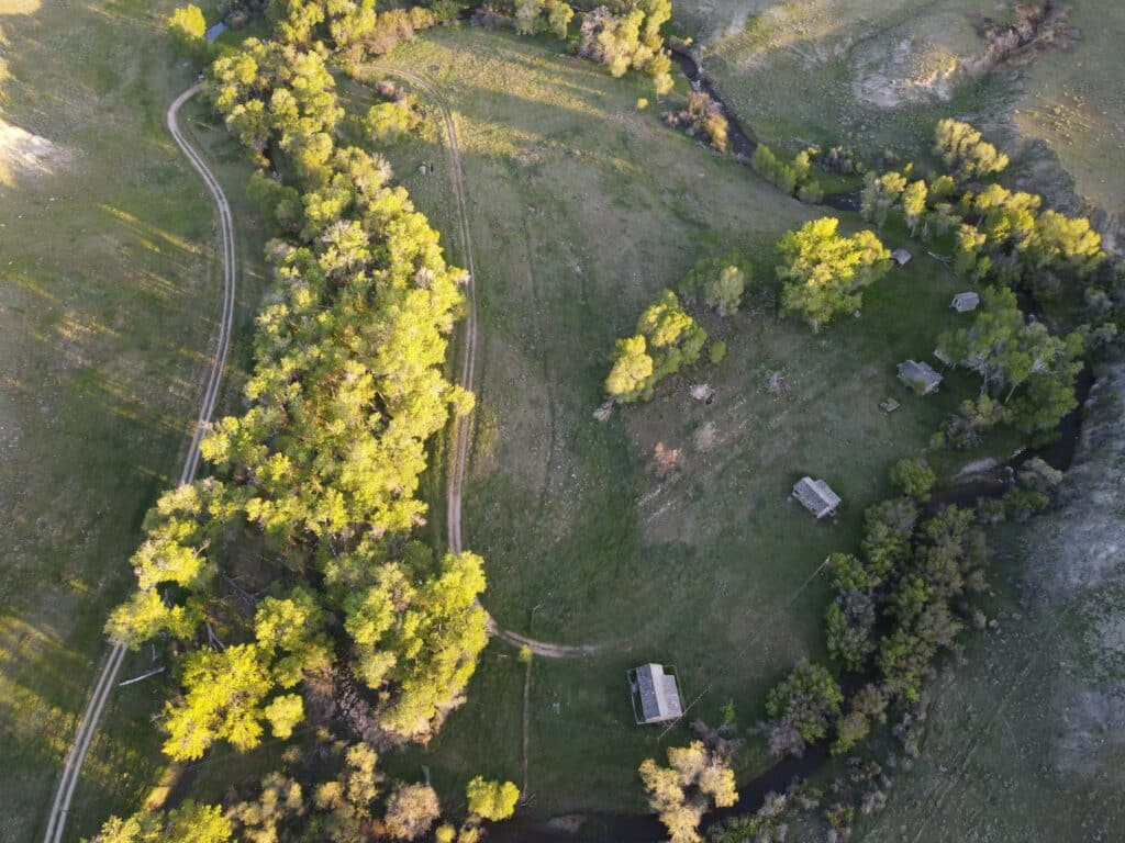 Aerial view of recreational land with winding dirt roads, clusters of green trees, small scattered cabins, and a stream curving through grassy fields under soft sunlight—perfect land for sale.