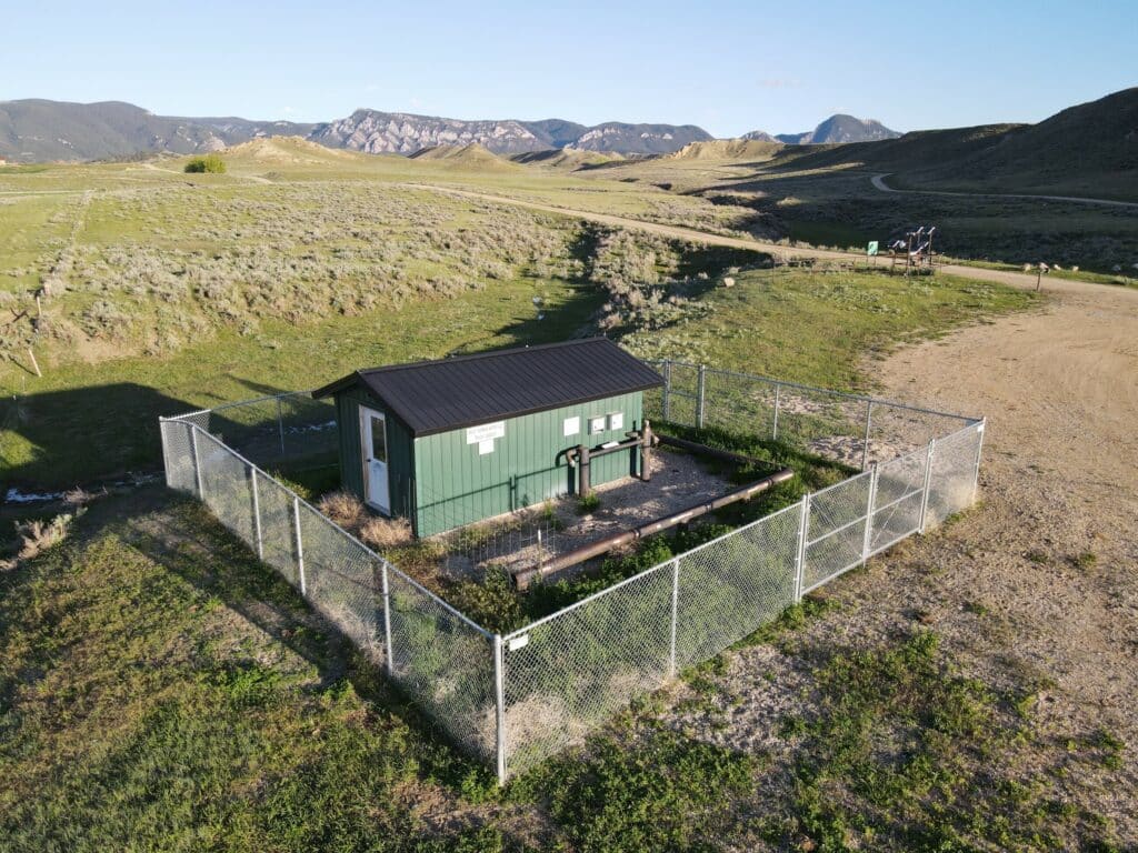 A small green building with a black roof sits inside a fenced enclosure on a grassy plain—perfect for a hunting property or ranch for sale—with mountains and rolling hills in the background under a clear sky.
