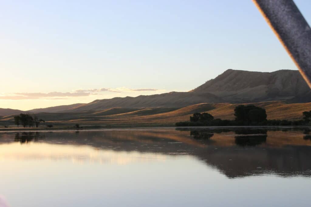 A calm lake reflects distant rolling hills and a large mountain at sunset, with soft golden light and a clear sky above—perfect scenery for experiencing recreational land or seeking an ideal ranch for sale.