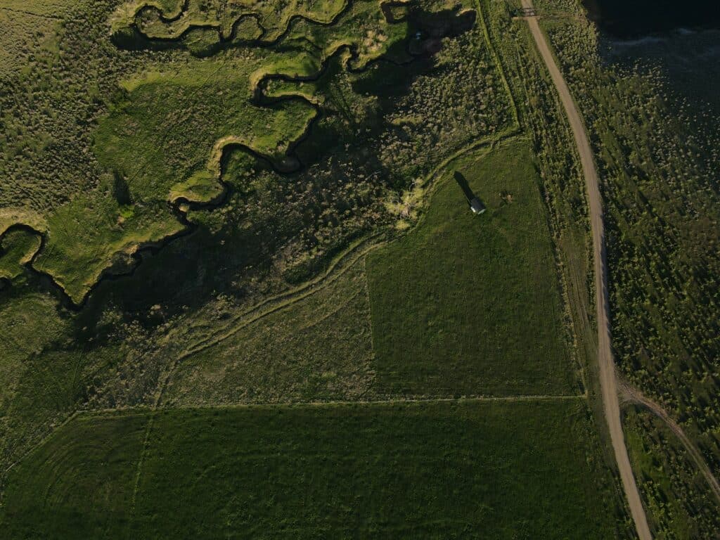 Aerial view of a green landscape with winding creek, rectangular grassy fields, a small structure casting a shadow, and a dirt road along the right—ideal ranch for sale or versatile recreational land.