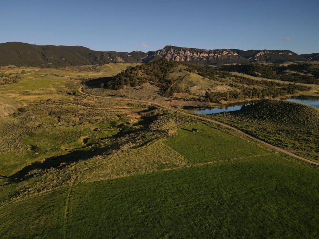 A wide landscape view of grassy fields, rolling hills, a winding road, a pond, and distant rocky cliffs under a clear blue sky—an ideal cattle ranch or hunting property in the late afternoon.
