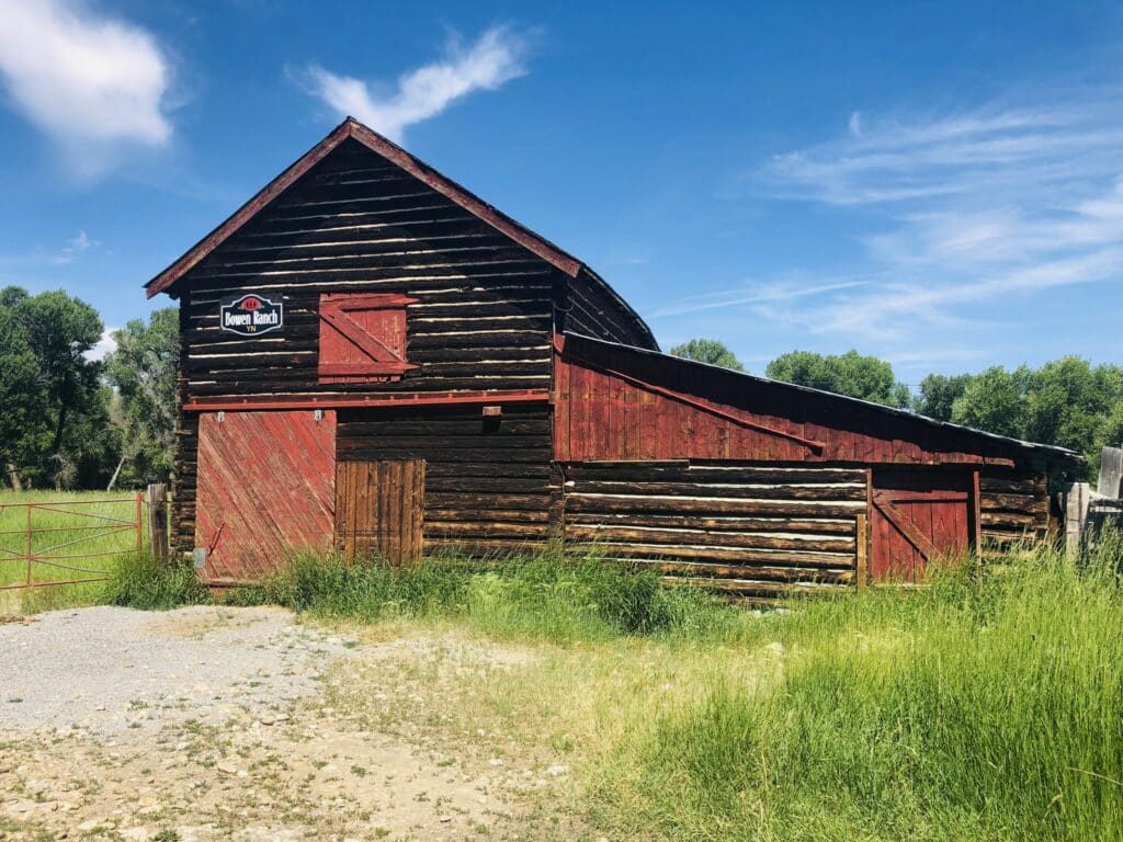 A rustic wooden barn with red doors and trim stands in a grassy field under a bright blue sky with scattered clouds. Ideal for a cattle ranch or hunting property, the land boasts trees and a metal gate in the background.