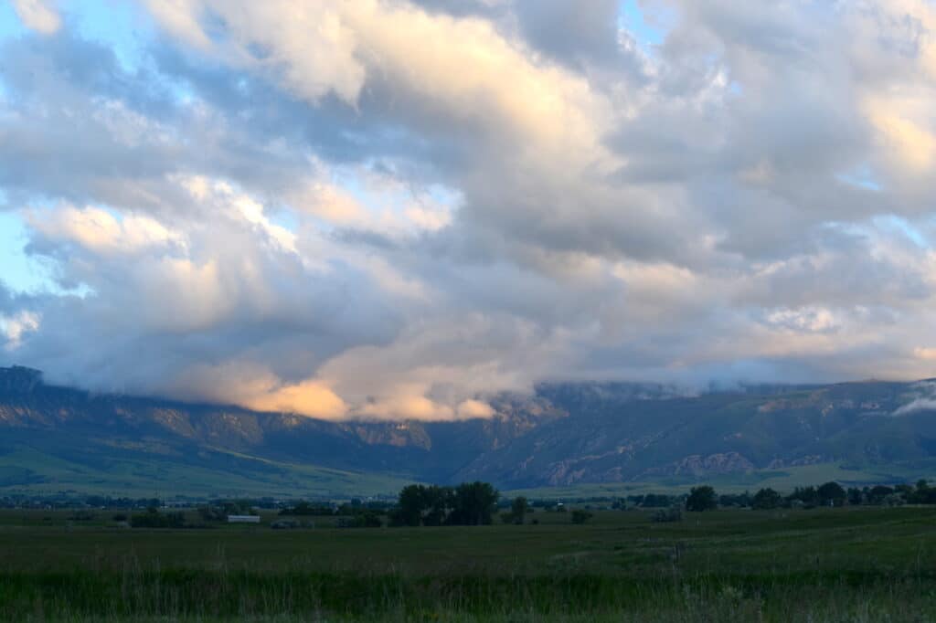 Clouds hang low over distant mountains at sunset, casting golden and blue hues. The foreground features recreational land with scattered trees and small buildings, making it an ideal hunting property under a dramatic sky.