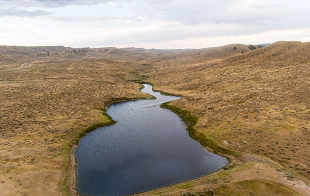 A small, winding lake surrounded by dry, rolling hills and sparse vegetation under a cloudy sky—perfect recreational land or potential hunting property.