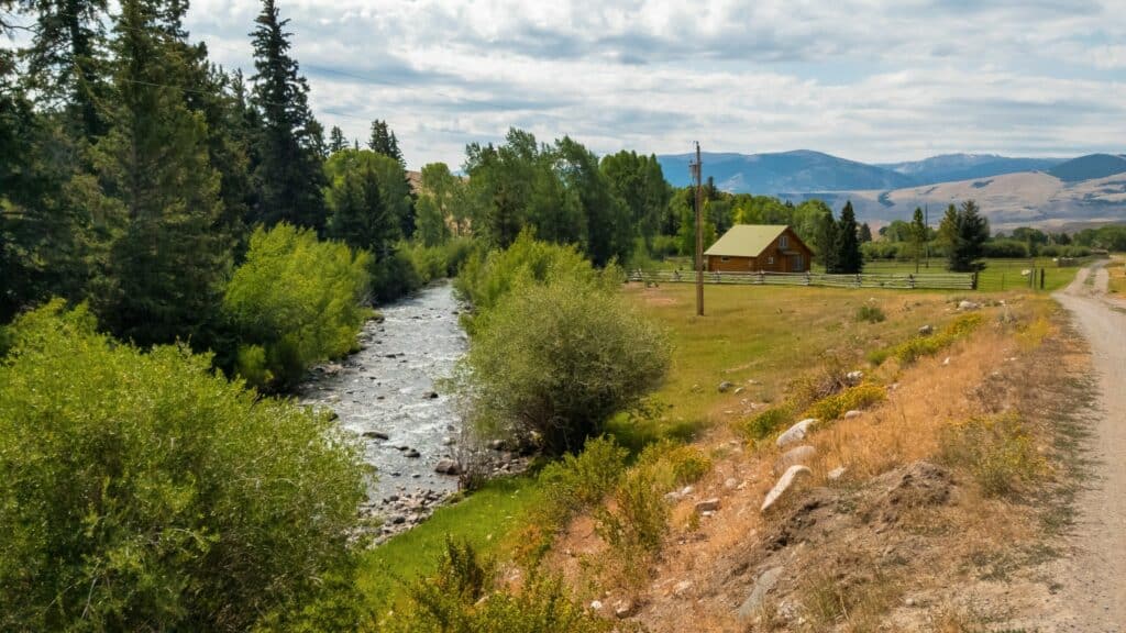 A small river flows beside a grassy field with a rustic cabin and wooden fence, surrounded by trees and mountains under a partly cloudy sky. This scenic ranch for sale features a dirt road running along the right side of the image.