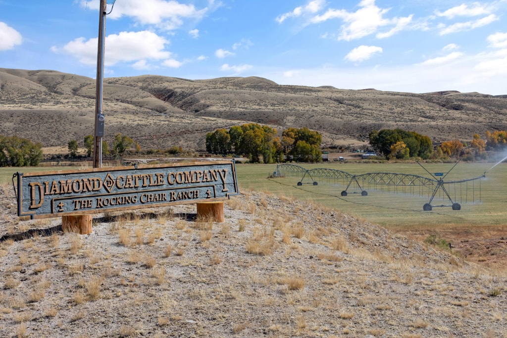 A wooden sign reading “Diamond Cattle Company, The Rocking Chair Ranch” stands on dry ground near rolling hills, green pasture, and a large irrigation system spraying water—a picturesque cattle ranch with trees and blue sky in the background.