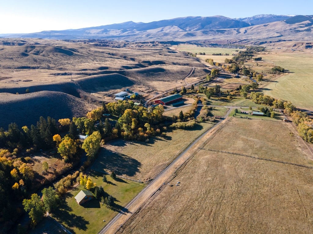 Aerial view of a rural landscape with scattered houses, trees, fields, and mountains under a clear blue sky. Dirt roads and farmland highlight this peaceful open-country scene—ideal for recreational land or a cattle ranch.