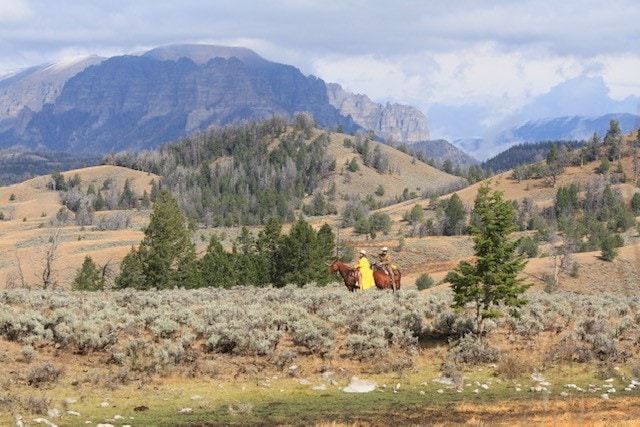 A person in a yellow jacket rides a horse through a sagebrush-filled landscape of rolling hills and scattered trees, with large mountains in the background—a classic scene on a hunting property or cattle ranch under a cloudy sky.