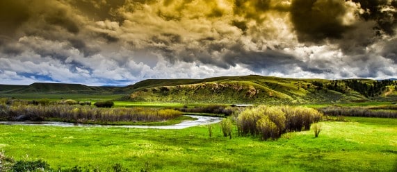 A dramatic sky with dark, swirling clouds looms over a lush green recreational land with rolling hills, a winding river, and dense bushes in the foreground.