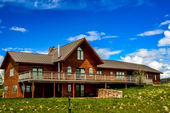 A large wooden house with a metal roof and wraparound deck sits on a grassy hill under a bright blue sky, perfect as a cattle ranch or hunting property.