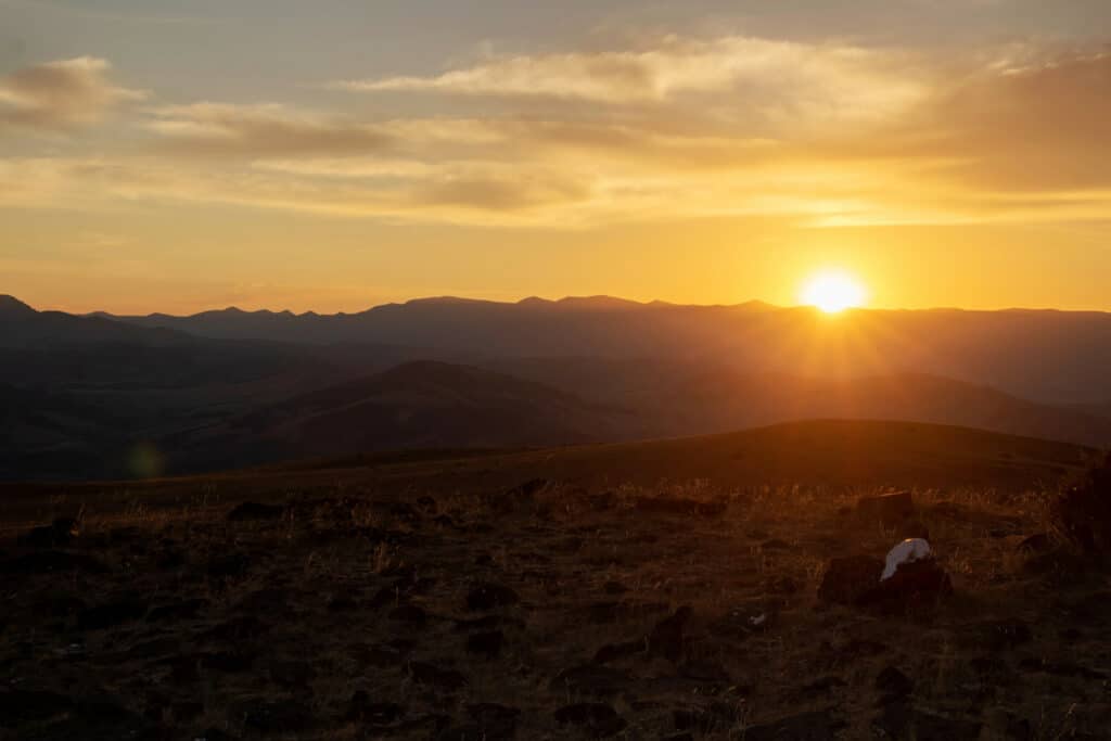 A golden sunset over distant mountain ranges, with the sun partially hidden behind the peaks. The foreground features rocky, grassy terrain—perfect recreational land for sale—under a glowing sky with scattered clouds.