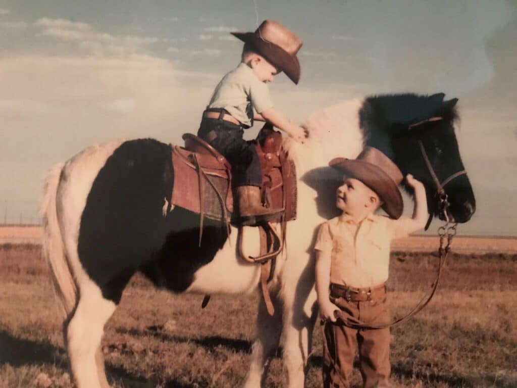 Two young children dressed as cowboys, one sitting on a black-and-white horse and the other standing beside it, outdoors on a grassy field under a blue sky, capture the spirit of life on a cattle ranch.