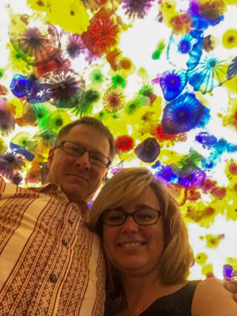 A smiling man and woman wearing glasses pose together beneath a vibrant ceiling covered with colorful glass art, reminiscent of the unique character you'll find on a ranch for sale or recreational land.