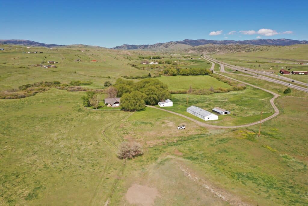 Aerial view of a rural ranch for sale with several buildings, surrounded by trees and open fields near a highway and rolling hills under a clear blue sky.