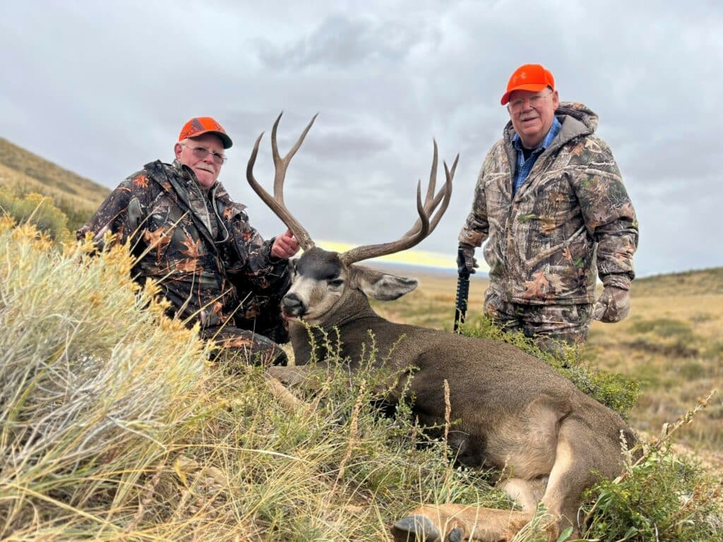 Two hunters in camouflage and orange hats pose with a large deer on the ground in a grassy, open landscape under a cloudy sky, showcasing the natural beauty often found on land for sale or a cattle ranch.