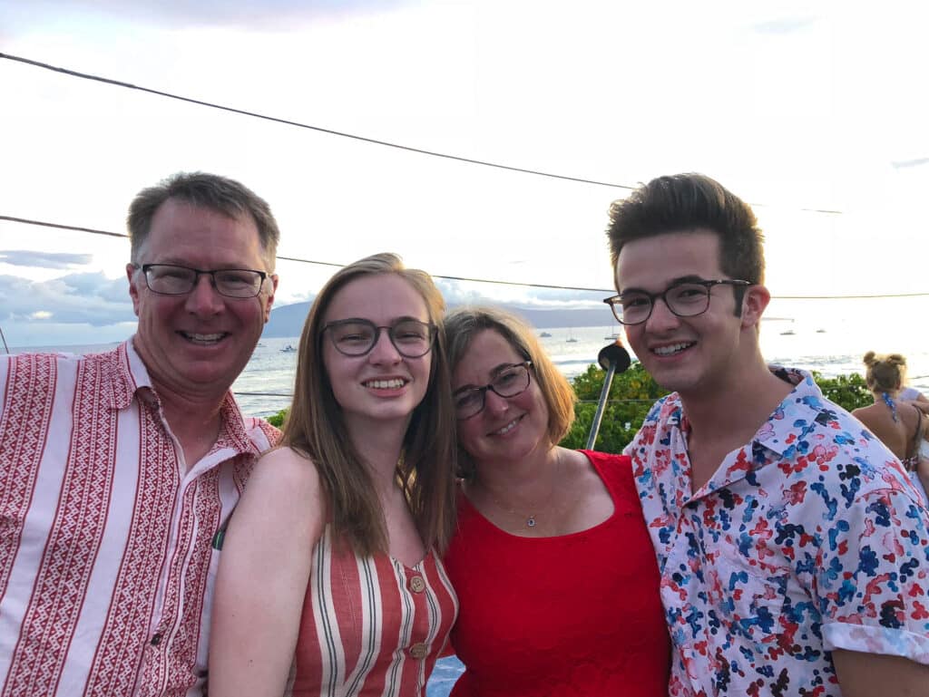 Four people smile and pose together outdoors near the beach at sunset. The group, standing on recreational land, includes two adults and two young adults in summer clothes with red, white, and pink patterns. Ocean waves and boats form a scenic background.