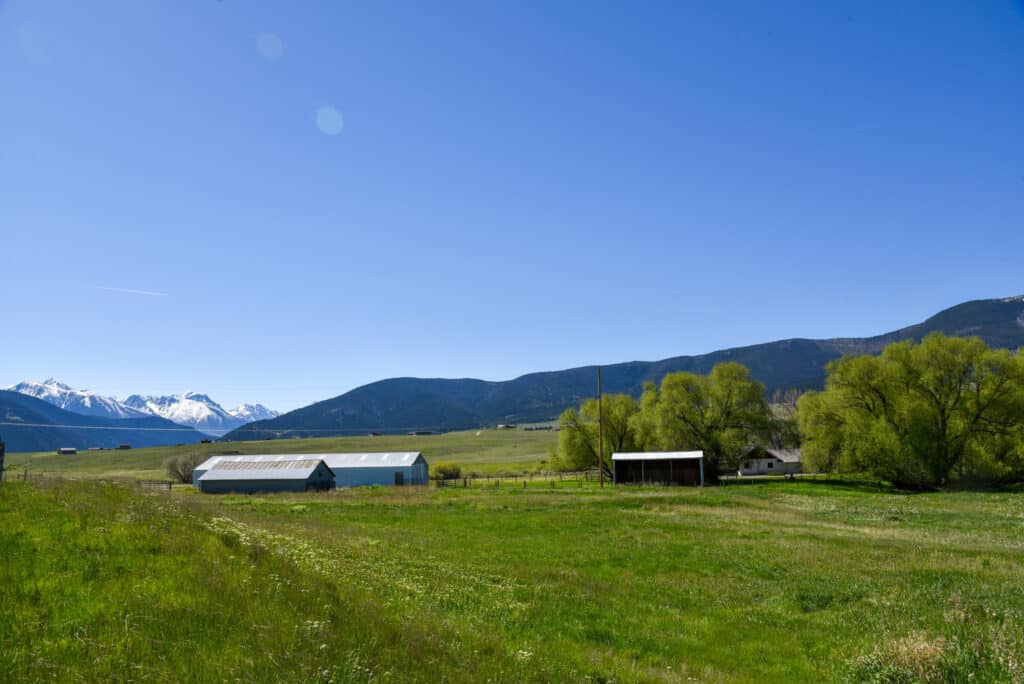 A rural landscape with green fields, a barn, a shed, a few trees, and snow-capped mountains in the distance under a clear blue sky—perfect hunting property or land for sale.